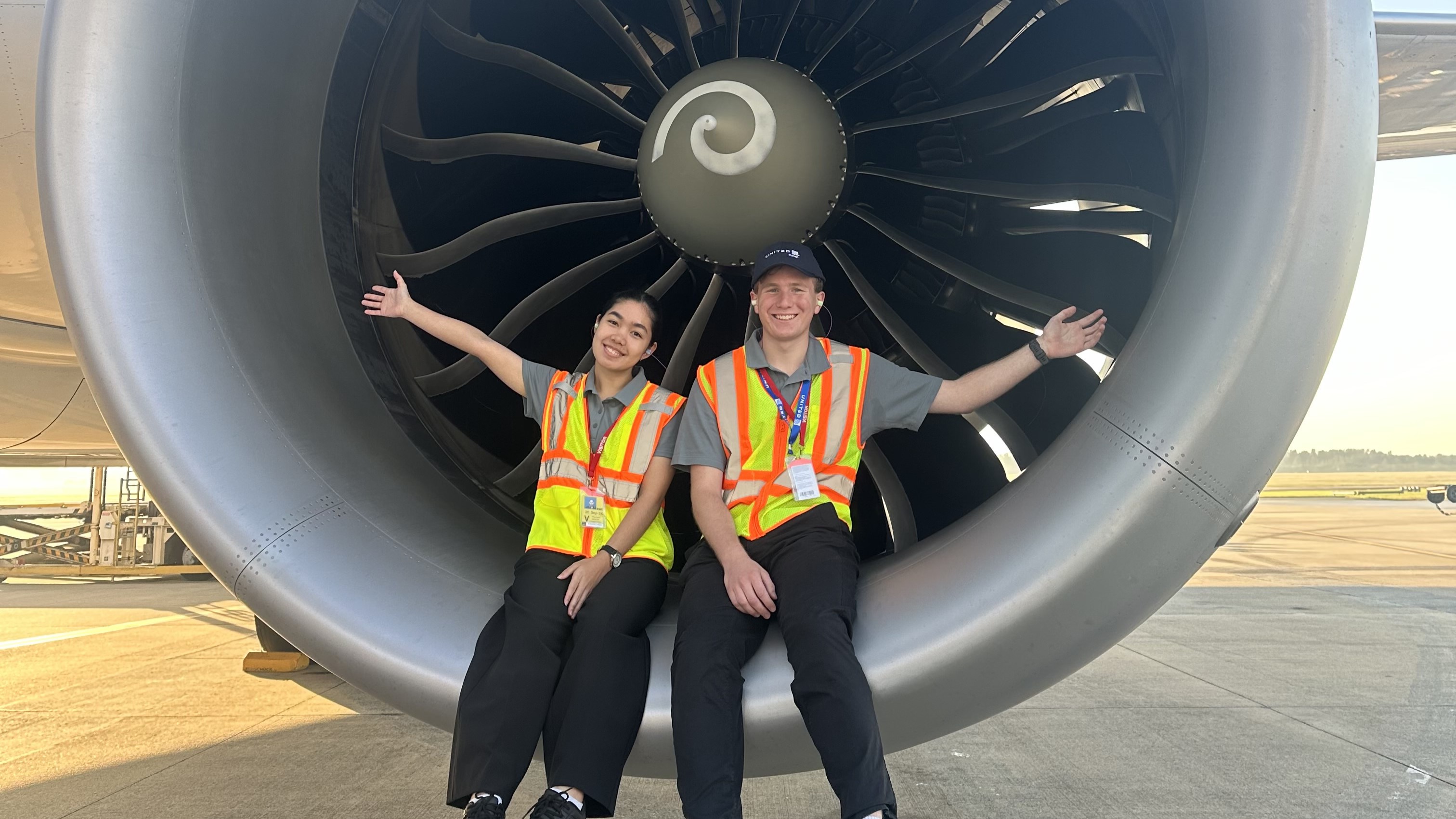 Two students sitting posing for a photo with an aircraft engine